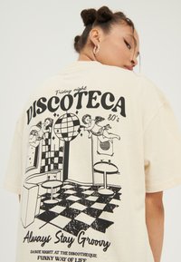 T-shirt oversize in cotone color crema con un design grafico nero che rappresenta una scena disco, pavimento a scacchi e elementi testuali legati agli anni '80.