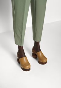 Sabots en cuir marron avec des bouts arrondis, talons en bois, accentués de petites rivets métalliques, associés à un pantalon ajusté vert clair.