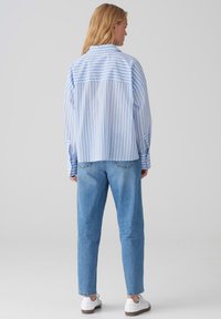 Camicia a righe blu e bianche realizzata in tessuto leggero, con colletto classico e vestibilità ampia, abbinata a jeans chiari in denim e scarpe bianche.