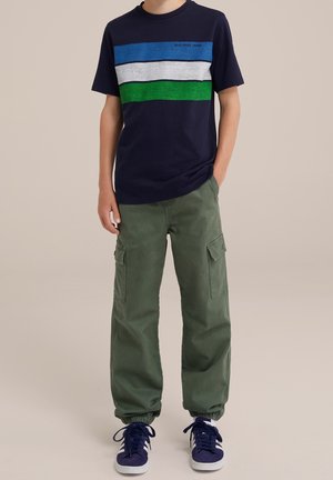 Niño con camiseta azul marino con rayas azules, blancas y verdes, pantalones cargo verde oliva con puños, y zapatillas azul marino con rayas blancas.