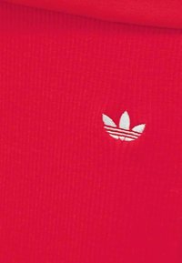 Tissu rouge présentant une texture côtelée subtile, avec un petit logo brodé blanc représentant trois formes de feuilles.