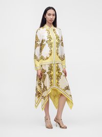 Frau trägt eine gelb-weiß gemusterte Bluse mit langen Ärmeln, einen asymmetrischen passenden Rock und goldene Sandalen mit hohen Absätzen.