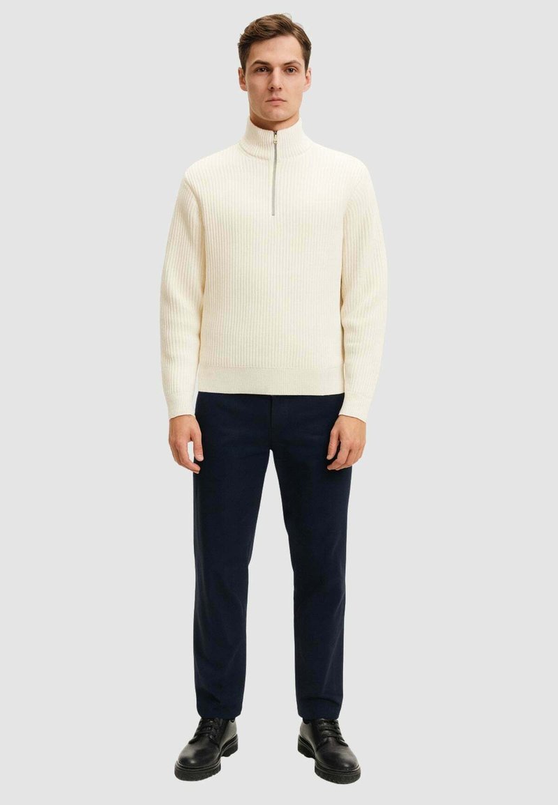 Pullover a coste color crema con colletto alto e mezzo zip frontale, abbinato a pantaloni blu scuro e scarpe nere.