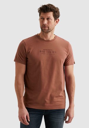 Uomo con capelli corti castani che indossa una t-shirt PME Legend color ruggine e jeans blu scuro, che guarda leggermente a sinistra.