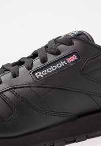Sapatilha preta de couro com superfície texturizada, design de atacadores, logotipo da Reebok e um pequeno detalhe da bandeira da União na lateral. Sola de borracha plana.