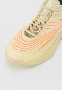 Baskets de sport beige avec des bandes courbes orange, une languette en mesh et des lacets beiges sur un fond clair.