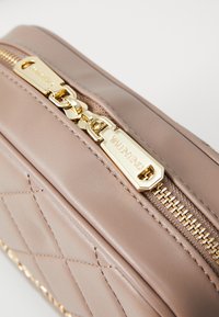 Artimas beigiško šonu suklijuoto odos maišo užtrauktukas su auksinėmis traukiklio detalėmis, įspaustais žodžiu "VALENTINO" šviesiame fone.