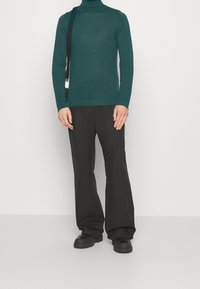 Tiefgrüner Rollkragenpullover kombiniert mit locker sitzenden schwarzen Hosen. Das Outfit zeichnet sich durch ein minimalistisches Design und eine glatte Textur aus. Schwarze Schuhe runden den Look ab.