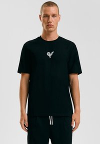 Zwarte katoenen t-shirt met een kenmerkend wit logo op de borst. Ronde halslijn en korte mouwen zorgen voor een ontspannen pasvorm.