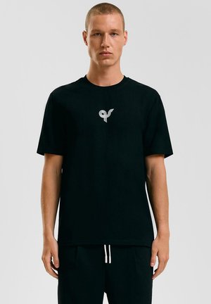 T-shirt en coton noir avec un logo blanc distinctif sur la poitrine. Encolure ras du cou et manches courtes pour un ajustement décontracté.
