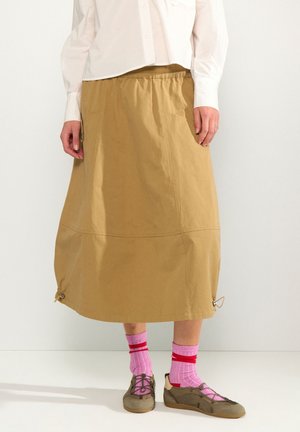 Personne portant une jupe midi beige avec des cordons au bas, des chaussettes roses avec des rayures rouges, des chaussures marron à lacets et une chemise blanche boutonnée.