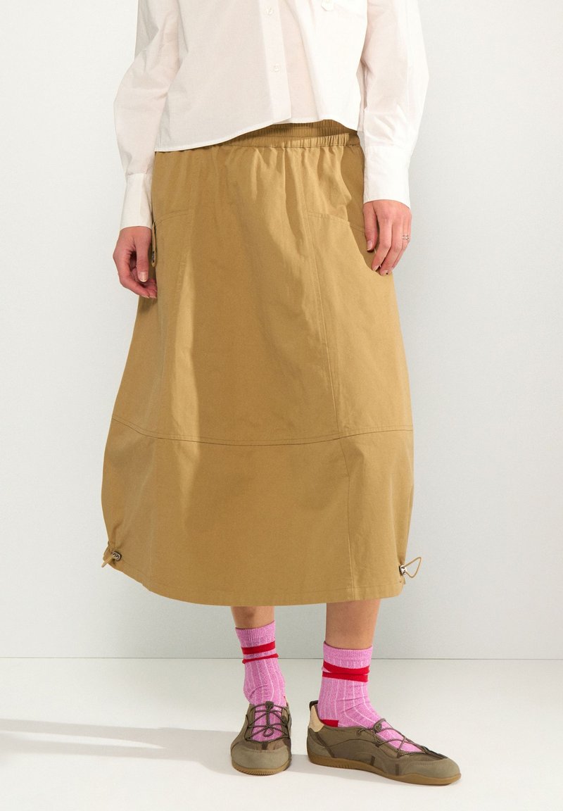 Personne portant une jupe midi beige avec des cordons au bas, des chaussettes roses avec des rayures rouges, des chaussures marron à lacets et une chemise blanche boutonnée.