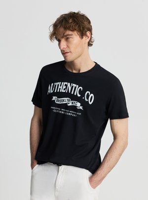 Młody mężczyzna z kręconymi włosami, ubrany w czarną koszulkę "Authentic.Co Brooklyn NYC" i białe spodnie, z rękami w kieszeniach, patrzący w bok.