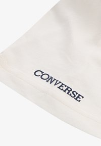 Tissu en coton couleur crème avec une texture lisse, présentant un logo "CONVERSE" bleu marine bien visible près de l'ourlet. Design simple et minimaliste.