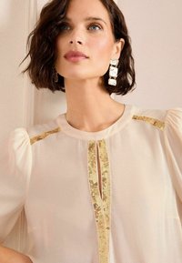 Blusa di crema chiara con maniche a palloncino, caratterizzata da dettagli in paillettes dorate sulle spalle e uno scollo a chiave.