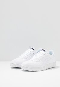Hvite joggesko med en teksturert skinnoverdel, snøresystem og fremtredende Asics-logo på tungen. Gummisåle med mønstrete sålett.