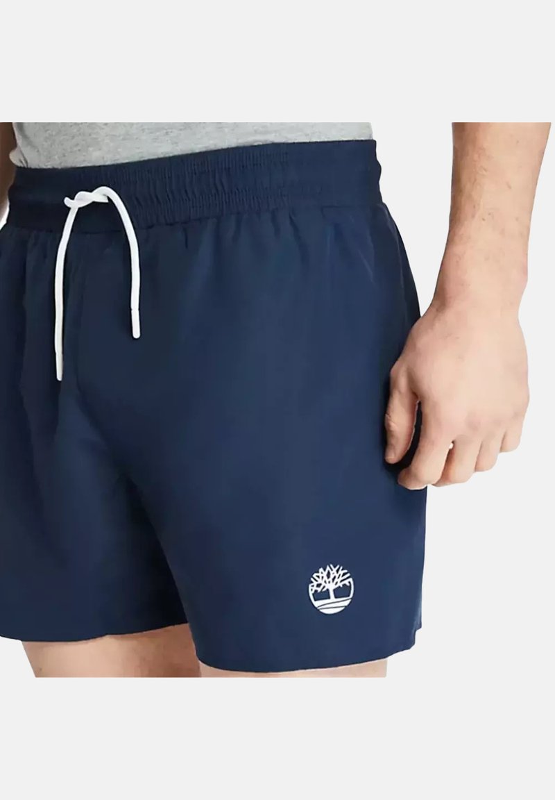 Pantaloni da bagno blu navy con coulisse bianca, vita elastica e un piccolo logo bianco a forma di albero sul lato sinistro inferiore. Tessuto leggero.