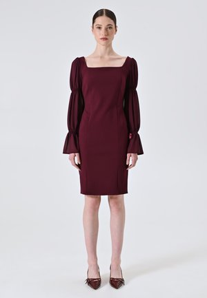 Bordeauxfarbendes, tailliertes Kleid mit quadratischem Halsausschnitt und langen Puffärmeln. Aus glattem Stoff, knielang, kombiniert mit bordeauxfarbenen spitzen Schuhen.