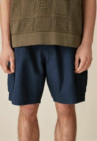 Olive green textured knit sweater with square patterns paired with navy blue shorts, featuring side pockets and a relaxed fit.