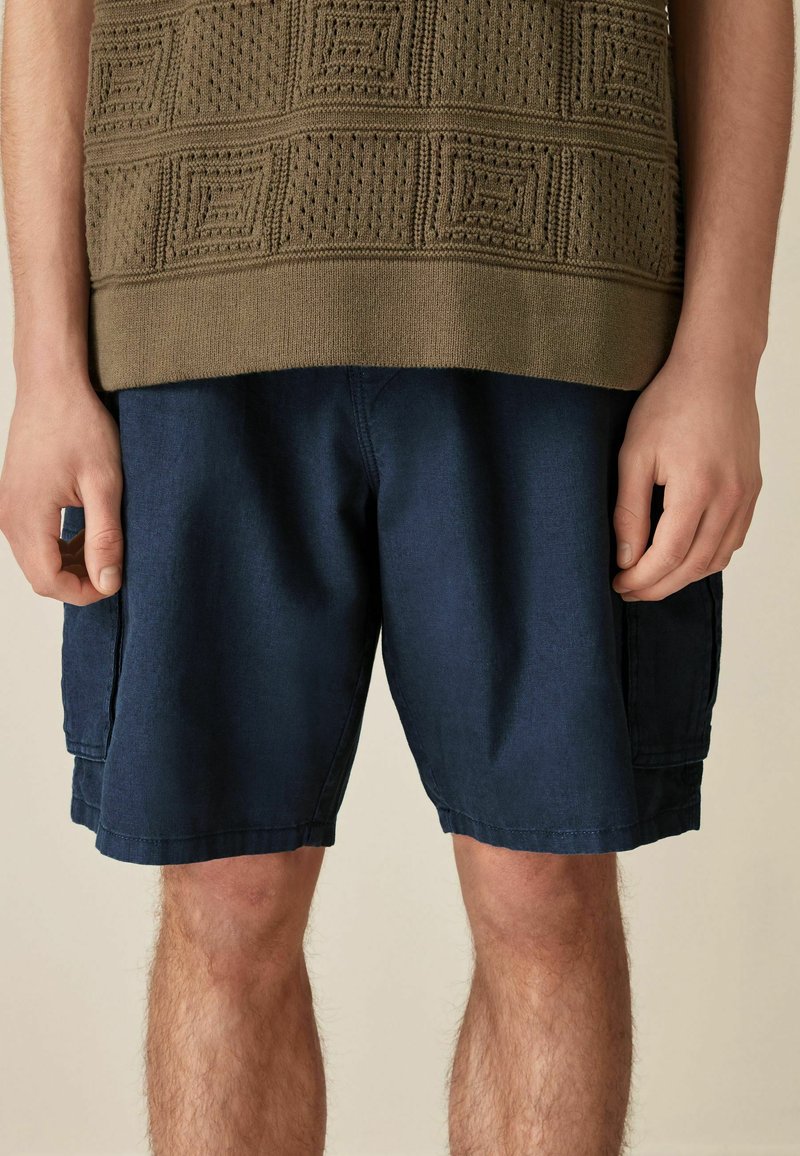 Olive green textured knit sweater with square patterns paired with navy blue shorts, featuring side pockets and a relaxed fit.