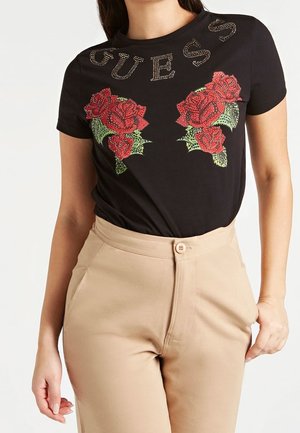 Sort T-shirt med korte ærmer med røde roseprint og "GUESS" i rhinsten, båret med beige højttaljede bukser.