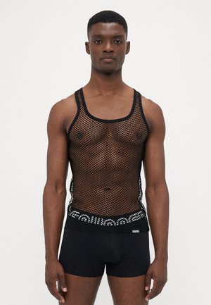 Mann in schwarzem Fischernetz-Top über gemusterten Unterhosen mit elastischem Bund und einfarbigen schwarzen Boxer-Shorts, stehend vor schlichten Hintergrund.