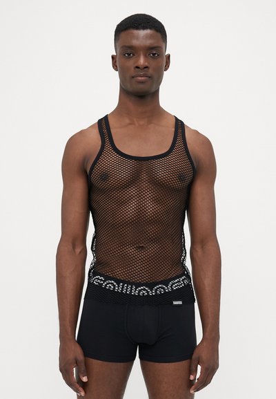 Mann in schwarzem Fischernetz-Top über gemusterten Unterhosen mit elastischem Bund und einfarbigen schwarzen Boxer-Shorts, stehend vor schlichten Hintergrund.