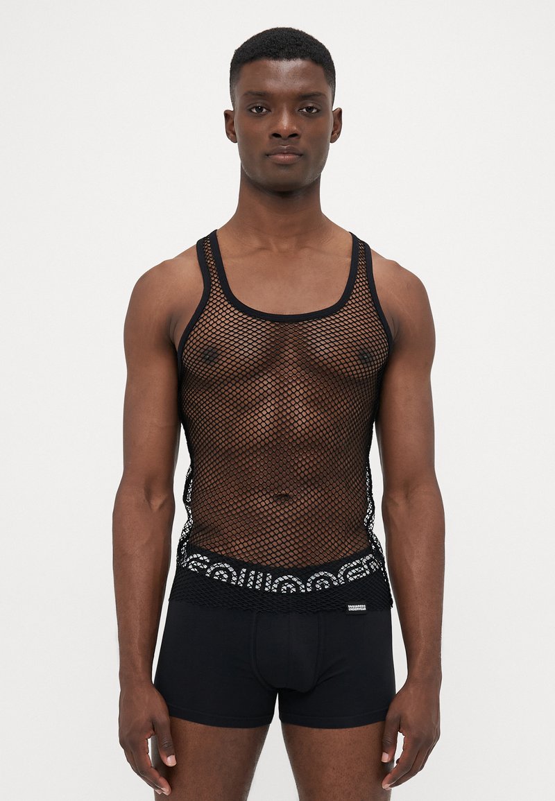 Mann in schwarzem Fischernetz-Top über gemusterten Unterhosen mit elastischem Bund und einfarbigen schwarzen Boxer-Shorts, stehend vor schlichten Hintergrund.