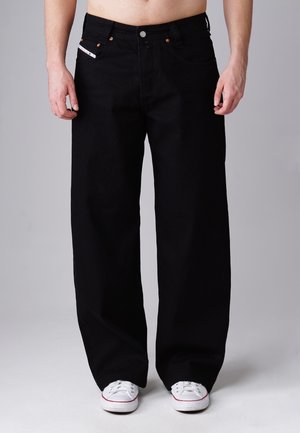 Male wearing loose black jeans and white sneakers, standing against a plain grey background, bare midriff visible.