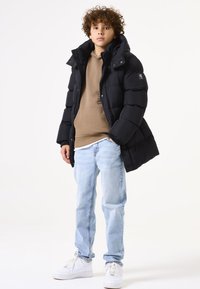 Schwarze gepolsterte Jacke mit hohem Kragen, kombiniert mit einem braunen Hoodie und hellblauen Jeans. Weiße Sneakers runden das lässige Outfit ab.