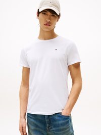 Junger Mann mit weißer Tommy Jeans Kappe, weißem, taillierten T-Shirt mit kleinem Logo und blauen Jeans, steht da mit einer Hand in der Tasche.