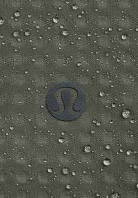 Waterafstotende groene stof met een gestructureerd patroon, voorzien van een zwart logo dat centraal is geplaatst en met waterdruppels verspreid over het oppervlak.