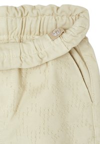 Beige cotton trousers with a gathered waistband, featuring a button detail and subtle textured pattern across the fabric.