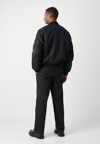Schwarze Bomberjacke mit gestepptem Muster, geripptem Kragen und Bündchen; kombiniert mit schwarzen Hosen und schwarzen Schuhen, von hinten betrachtet.