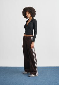 Μαύρο μακρυμάνικο τοπ με φερμουάρ, καφέ παντελόνι με φαρδιά μπατζάκια και άσπρες ρίγες στα πλαϊνά, και γκρι ριγέ παπούτσια slip-on σε μπλε χαλί φόντο.