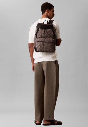Jeune homme portant une chemise à manches courtes de couleur claire, un pantalon marron, des sandales noires et un sac à dos marron et noir à motifs, debout devant un fond gris.