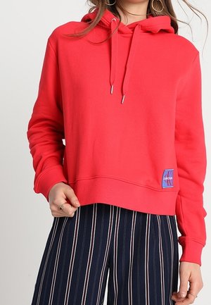 Czerwona bluza z kapturem ze ściągaczami i małą naszywką z logo, noszona z granatowymi pionowo prążkowanymi spodniami.