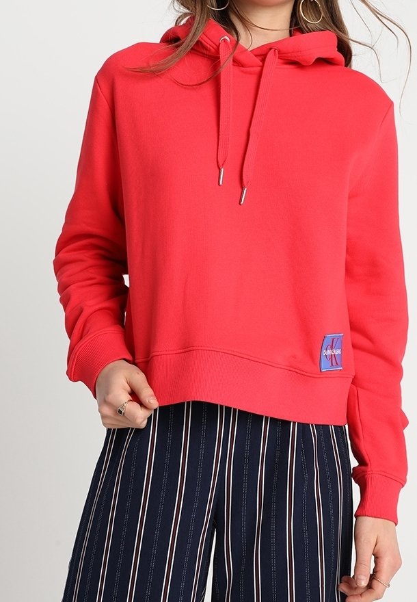 Sweat à capuche rouge avec cordons et petit patch logo, porté avec un pantalon bleu marine à rayures verticales.