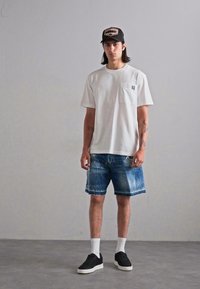 T-shirt bianca a maniche corte con taschino, pantaloni corti in denim blu con lavaggio sbiadito, sneakers nere con suole bianche e berretto nero con logo.