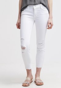 Vaqueros blancos desgastados con roturas en las rodillas, corte slim, cintura media y dobladillo deshilachado. Combinados con sandalias planas en beige y plata.