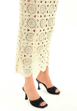 Mules à talons hauts noirs avec un dessus texturé, design à bout ouvert, associées à une jupe en crochet couleur crème avec des motifs circulaires.