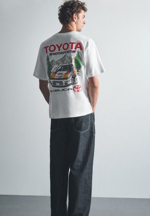 Homme portant un t-shirt blanc Toyota Motorsports Celica avec un graphique de voiture, montrant son dos, debout devant un fond clair uni.