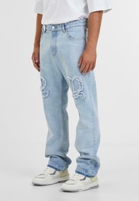 Jean en denim déchiré bleu clair avec des accents effilochés et une coupe décontractée, assortis à des baskets blanches avec des détails gris clair et jaunes.