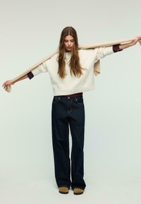 Cream-colored, chunky knit sweater, cropped at the waist. Dark blue wide-leg jeans, visibly textured. Model holds a beige scarf.