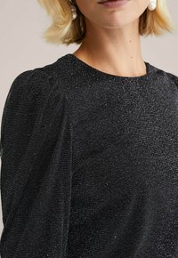 Sort tekstureret top med en glitrende overflade, der har pufskuldre og en rund halsudskæring. Stoffet har en subtil metallisk glans.