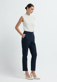 Valkoinen hihaton paita, jossa on tikkulaitakuformaatti, yhdistettynä tummansinisiin räätälöityihin housuihin. Mallilla on päällään nudet korkokengät ja kultaketjut.