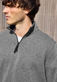 Šedá mikina na zip s vysokým límcem, vyznačující se hladkým povrchem a černým zipem na rozmazaném dřevěném pozadí.