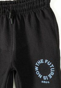 Pantalon de survêtement noir avec ceinture élastique et cordons, arborant un texte circulaire bleu "THE FUTURE IS NOW" ainsi que de petits caractères japonais en dessous.