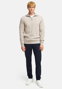 Helle beige Strickjacke mit einem halben Reißverschluss am Kragen, gerippten Bündchen, kombiniert mit navyfarbene Hosen und weißen Sneakern. Einfaches, strukturiertes Design.