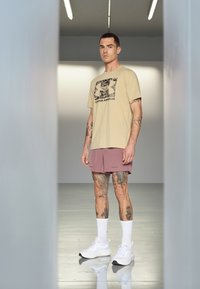 T-shirt bege de mangas curtas com uma estampa gráfica, combinada com calções cor-de-rosa claros. Ténis brancos desportivos e meias brancas até ao joelho.